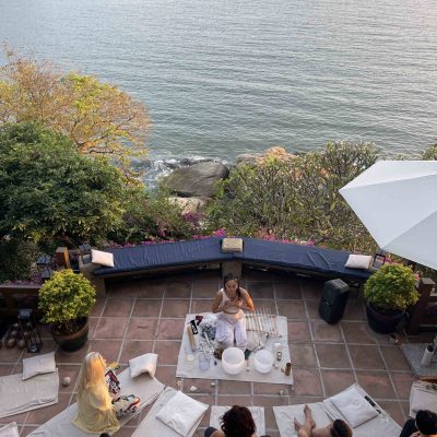 Participants lying down for a sound bath under a large thatched roof gazebo with a practitioner playing singing bowls.